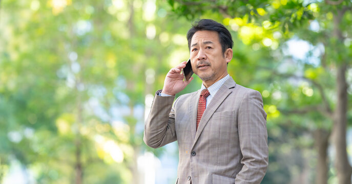 新緑に立つスーツを着たミドル男性ビジネスマン　スマートフォン　スマホ