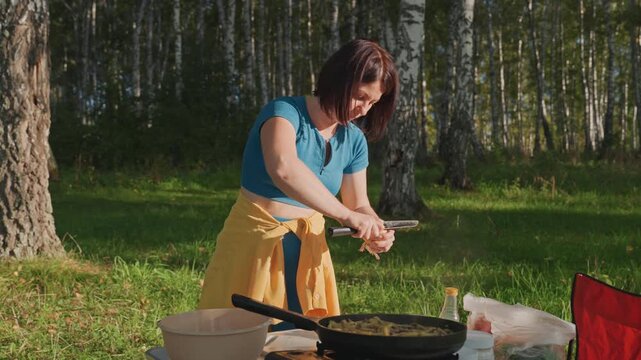 Caucasian woman camping prepping eggs birch forest, sunlit clearing with portable stove on folding table, red camp chair and black dog nearby, focused hands cracking shells into bowl, rustic summer