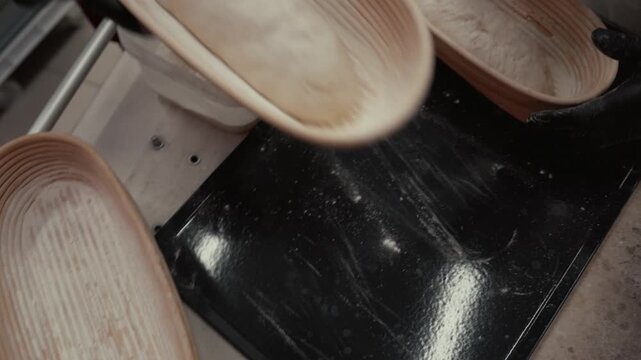 Baker in black gloves lifts dough from proofing basket onto floured surface, preparing for shaping and baking, with additional dough pieces visible on the tray