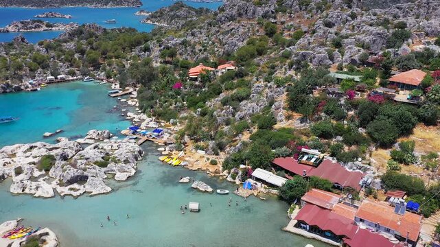 Aerial flyover of Kekova island shoreline revealing turquoise coves, rocky outcrops, waterfront restaurants and kayaks on a clear summer day, Antalya, Turkey