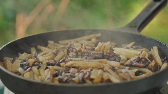 Sizzling fries and mushrooms in pan, hands stir with wooden spatula in castiron skillet, steam rising over golden crisp fries and sauteed mushrooms, smoky aroma fills campsite, cozy rustic camping
