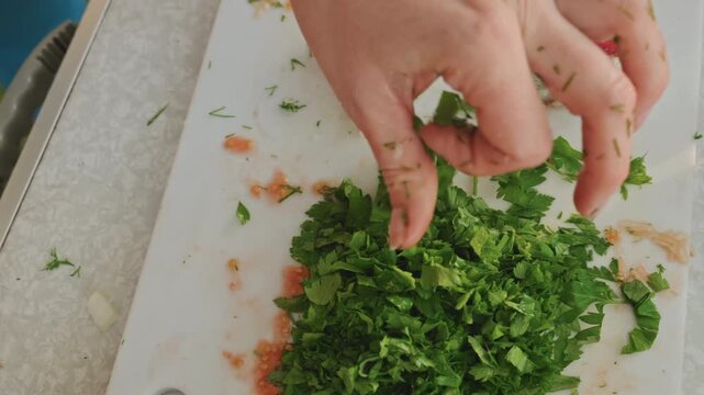 Closeup mincing parsley on board, outdoor chef shaping herb pile for garnish, small grater and clove visible, attentive finger placement and repeated knife strokes, campsite table atmosphere