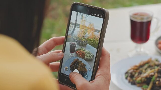 Phone closeup camping food photography showing woman hands framing plate and glass on portable table. Smartphone screen reflecting plated salad and drink, careful angle and filter preview visible,