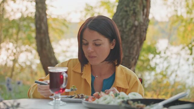 Caucasian woman camping lakeside enjoying warm meal and herbal tea at outdoor table, sunset glow, trees and calm water in background, peaceful solitude, savoring food with relaxed expression, skillet