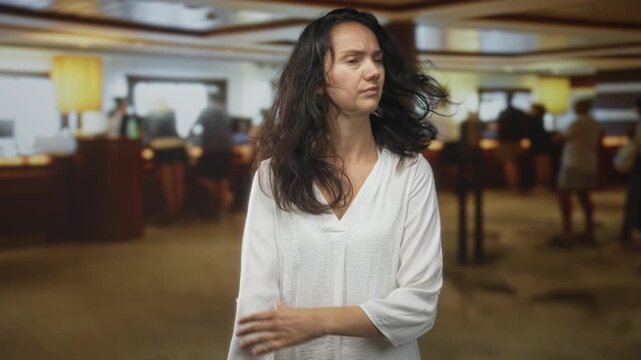 Woman rubbing forearm and rolling up sleeve at a reception counter in a building while glancing sideways, blurred queue behind her; discomfort travel.