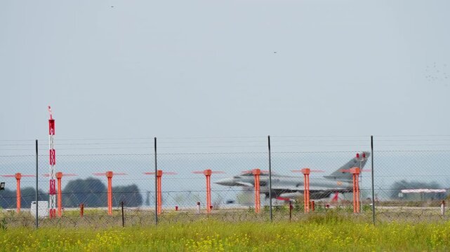 A grey military fighter jet is seen taxiing on an airport runway. The view is from behind a chain link security fence at a military airbase with a Zona Militar sign visible
