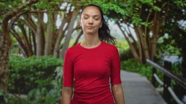 Woman waves hand with slight head tilt on a forest path beside a wooden railing, wearing red shirt and ponytail; indifference.