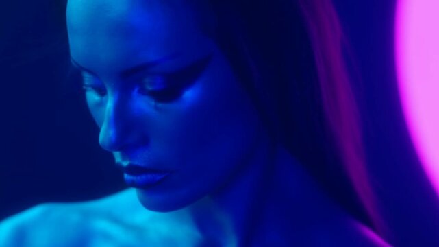 Slow motion close up of woman lowering head and tilting gaze in neon studio portrait with blue and pink rim lighting dramatic makeup intimate mood
