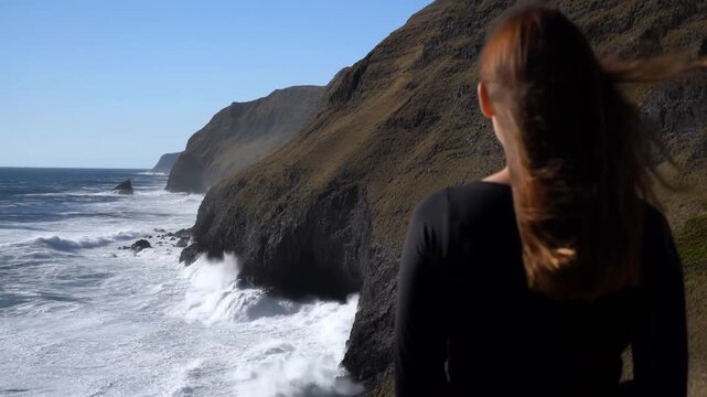 Tracking shot of woman standing and gazing at crashing waves along rocky cliff coastline with windblown hair during clear daytime