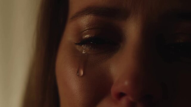 Slow motion close up of woman crying as tear rolls down cheek in dim warm indoor room portrait, intimate low light interior
