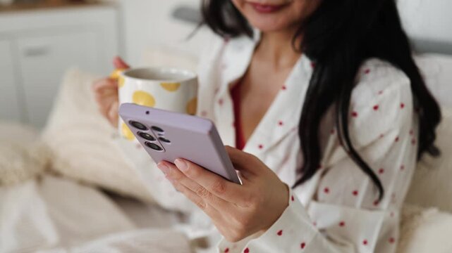 happy asian girl is smiling while having fun using chat apps to read and send text message to friend on the phone during morning coffee break on bed at home.