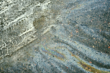 Naklejka premium Oil slick on rough asphalt road creating an iridescent rainbow pattern and showing white tire marks, indicating pollution