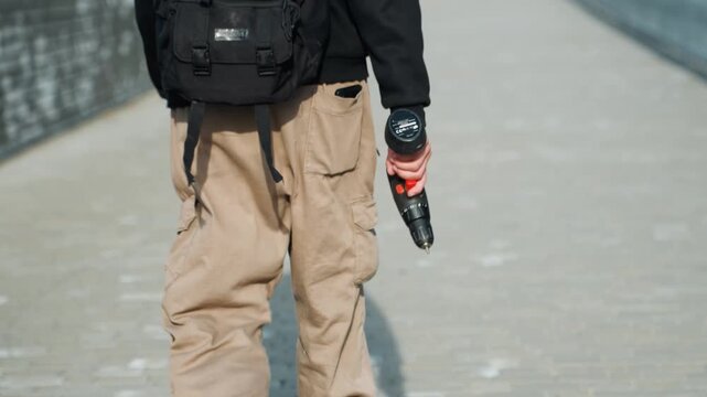 White man walking with drill along city bridge, tan cargo pants, black backpack, wristwatch on left hand, steady stride, tense atmosphere suggesting covert technician or suspect, daytime light