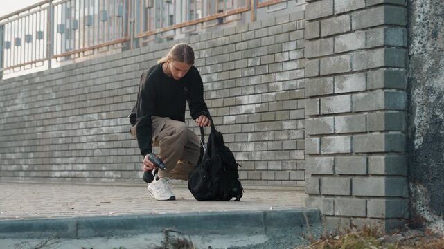 Urban courier white youth rummaging through backpack on brick walkway, pausing to crouch at curb, adjusting strap and checking contents, casual cargo pants and sneakers, muted city backdrop.