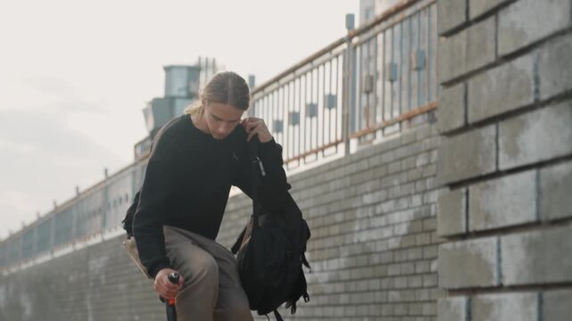 Caucasian man packing backpack on riverside bridge, checking cordless drill, overcast morning, brick wall backdrop, subdued palette, candid urban commute vibe, focused expression, hands sorting tool,