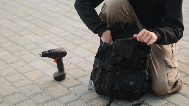 Young man retrieving drill from backpack, kneeling on pavement beside electric scooter, pulling toolkit and screwdriver, handson repair and battery check, practical maintenance scene, technician