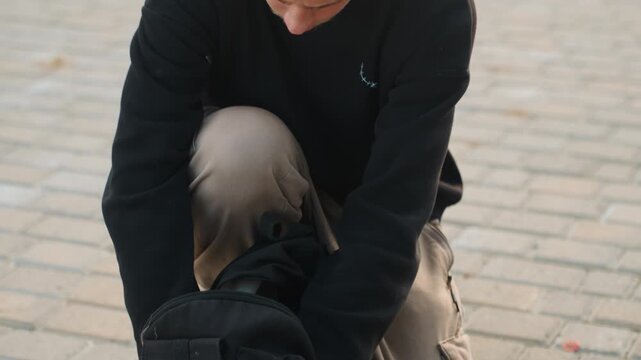 Young white man inspecting hammer next to backpack. Handyman checks chisel and hardware while kneeling on concrete, fingers steady, methodical selection and closeup hands create authentic urban craft