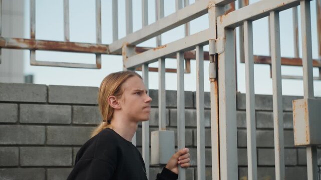 Young blonde examining rusted gate bars up close, steady grip on vertical rail, assessing fasteners and corrosion before using drill, determined expression, gritty urban contractor mood with cool
