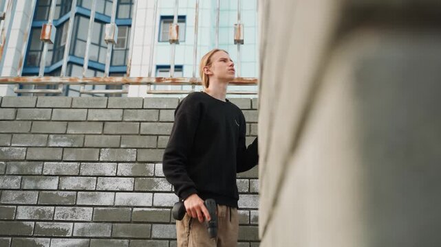 White man scouting brick wall, holding camera looking upward, urban stairway and concrete facade, cargo pants and black sweater, introspective mood and candid composition, testing angles and texture