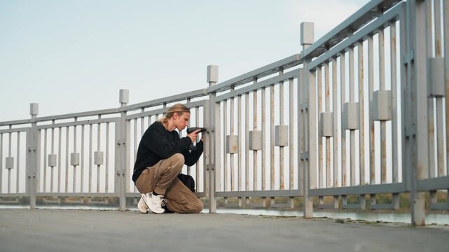 White inspector crouching by bridge railing checking bolts and metal condition, pragmatic assessment on waterfront platform, professional evaluation with tools and report, focused male performing