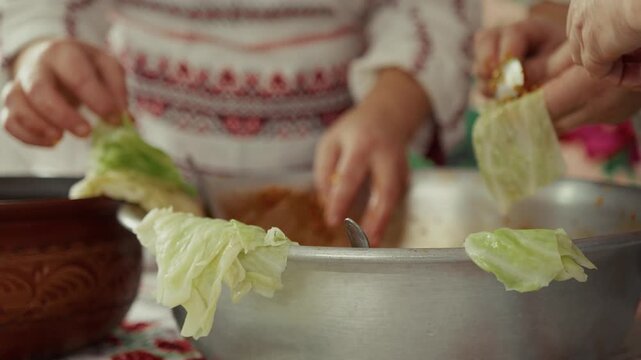 Hands of women in traditional attire prepare stuffed cabbage rolls using fresh cabbage leaves and a large bowl filled with filling on a colorful tablecloth in a bright kitchen
