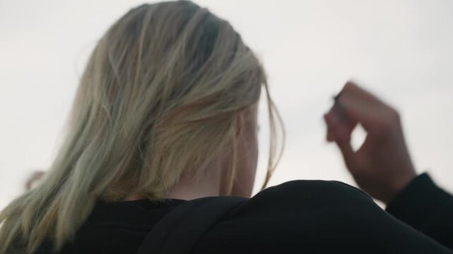 Blonde white woman back outdoors tying ponytail, hands smoothing strands, windblown texture, cloudy dusk, black hoodie, urban overpass, contemplative cinematic mood, intimate closeup, quiet solitude.