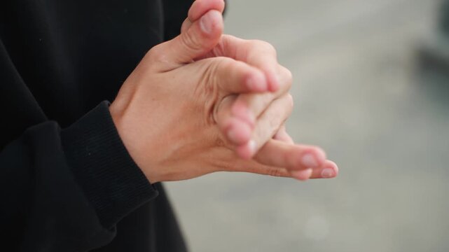 Close up hands rubbing knuckles outdoors, person in black sweater performing anxious hand motions, slow finger stretches, gentle palm rubs, candid urban backdrop, mood of preparation and focus, roles