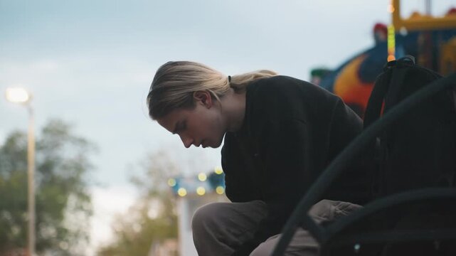 Young caucasian man sitting bench looking down backpack at shoulder and carnival lights bokeh behind, quiet reflective posture as he checks phone and smooths jacket student waiting for ride, traveler