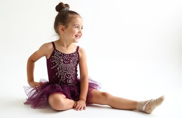 Naklejka premium Little girl in sparkly purple dance costume sits on floor. She wears ballet slippers and has her hair in a bun. Young dancer smiles while sitting down, preparing for her next move.