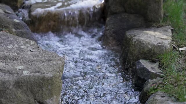 Clear water flows gently over smooth stones. Ripples splash softly against mossy rock surfaces. Nature's quiet movement creates soothing visual rhythm. Ideal for scenes needing calm, peaceful