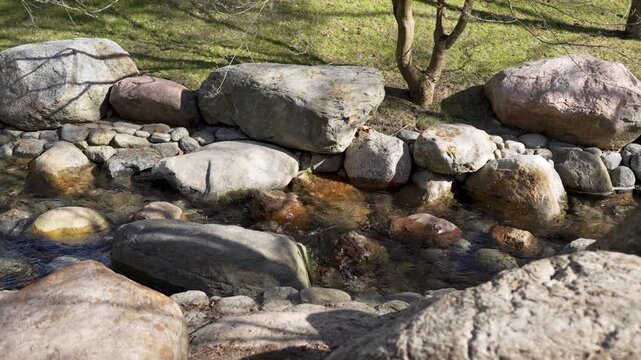 Calm stream flows gently over smooth stones. Rocks line the water's edge in earthy tones. Sunlight glints off ripples and mossy surfaces. Peaceful atmosphere invites quiet contemplation