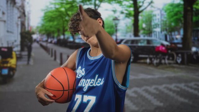 Teenage boy in blue los angeles jersey holding basketball points finger forward on city street with curly hair visible; confidence swagger.