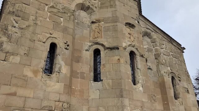 Ancient georgian orthodox stone church facade slowly tilting up revealing religious bas reliefs and narrow arched windows under a cloudy sky in a historical setting