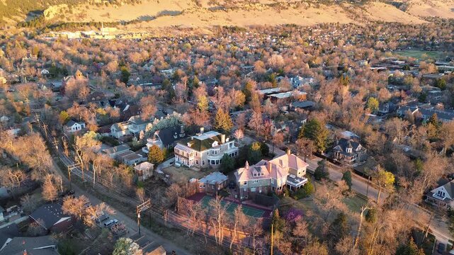 Upscale Boulder foothill neighborhood climbs gently toward open space, curving streets, large Victorian homes, tucked tennis court near hill base. Low warm light, sparse lawns, rocky slopes rise