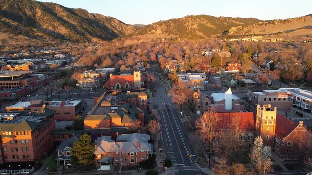 Downtown Boulder, Pearl Street corridor sunlit foothills, early spring trees Front Range. Warm golden light skims brick roofs, pale stucco walls, bare branches, patchy forested slopes ridgelines