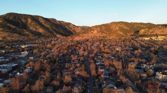 Northwest Boulder neighborhoods spread foothill ridge, winding streets, dense housing against mountain base. Long golden shadows trace rooftops, chimneys, bare branches, rugged hillside backdrop