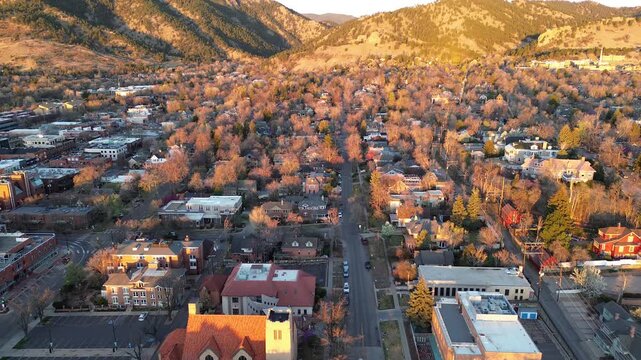 Central Boulder Street grid, church tower, low commercial blocks extend toward rising Front Range foothills. Brick facades, flat roofs, leafless treetops, rocky slopes layer distant ridgelines