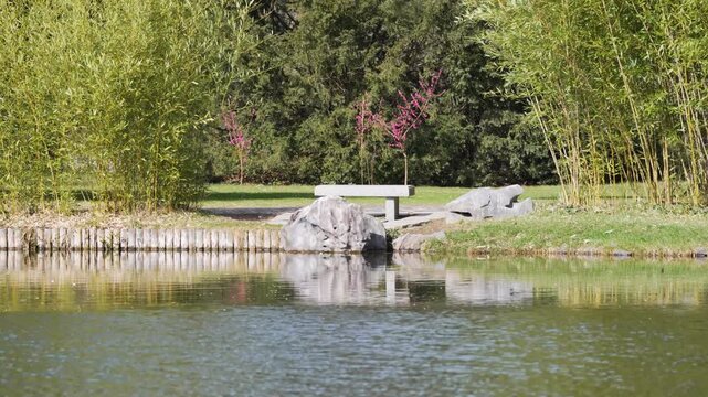 Still pond reflects green trees and a stone bench. Bamboo stalks sway gently beside the water. Pink blossoms peek from behind leafy branches. Serene atmosphere invites quiet contemplation