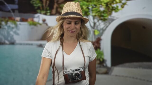 Woman tourist with vintage camera around her neck extends arm toward camera at hotel building pool area; joy vacation.
