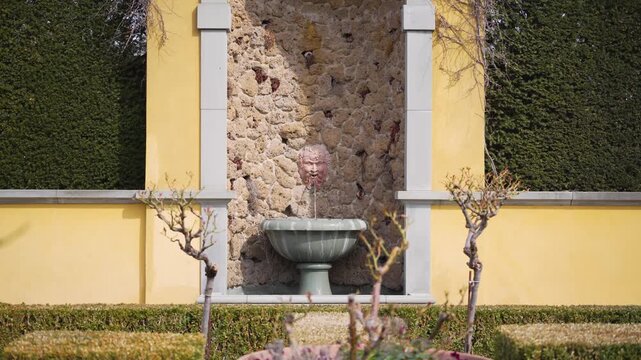 Sunlit garden fountain with stone basin. Yellow wall frames aged stone backdrop. Green hedges flank symmetrical structure. Tiny trees lean toward water's gentle flow. Ideal for tranquil, classical