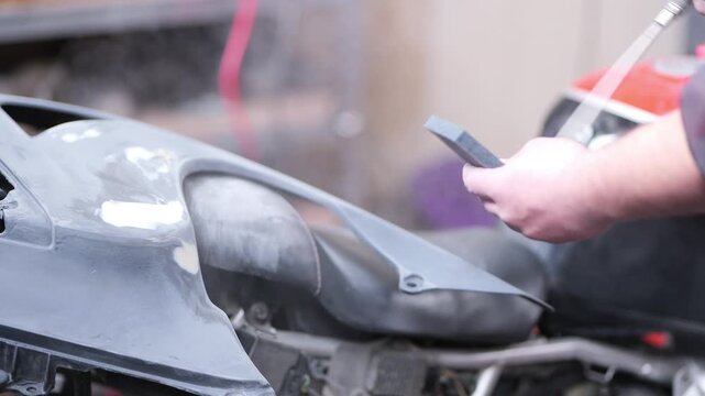 A detailed close-up video shows a mechanic painter using compressed air to blow dust off a sanded body filler surface, while cleaning a foam sanding block as fine particles disperse in the air, 2026
