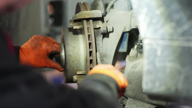 A close-up handheld video shows a mechanic wearing orange gloves cleaning brake components with a wire brush around the brake disc, removing rust and debris to ensure proper brake function, 2026