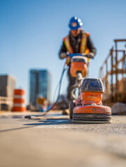 Fototapeta premium Construction worker operating a surface grinding machine on a concrete site with urban buildings and safety equipment visible in bright daylight