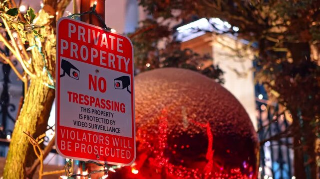 Private property warning sign with Christmas decorations in background night. Red metal sign with security camera icons in front of festive holiday lights and a large red bauble at night.