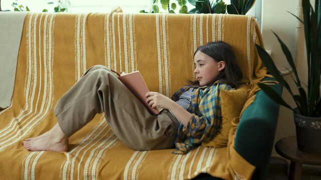 Happy preteen girl reading a book on a cozy couch at home, smiling preteen child enjoying literature in a bright living room, 4K high quality lifestyle footage.