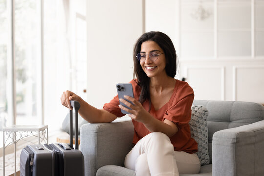 Smiling Brazilian woman using smartphone seated in armchair with suitcase while waits for taxi or airport transfer, prepare for flight, messaging through mobile app, make check-in, check boarding pass