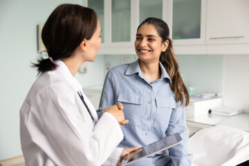Obraz premium Professional female GP doctor in white uniform holding digital tablet shaking hands with smiling Indian woman patient at hospital office during consultation. successful treatment, healthcare support