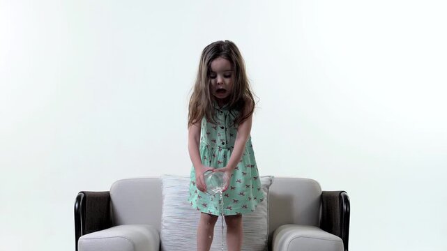 Little schoolgirl with vivid emotions of surprise spills a glass of water on a chair and the floor. Slow motion