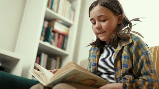 Happy preteen girl reading a book on a cozy couch at home, smiling preteen child enjoying literature in a bright living room, 4K high quality lifestyle footage.