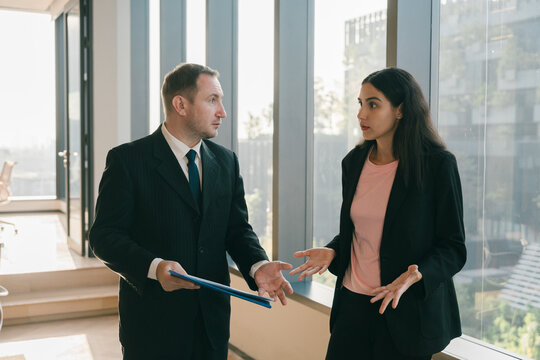 Businessman and businesswoman having serious discussion in office hallway with large windows, expressing concern and confusion during work conversation
